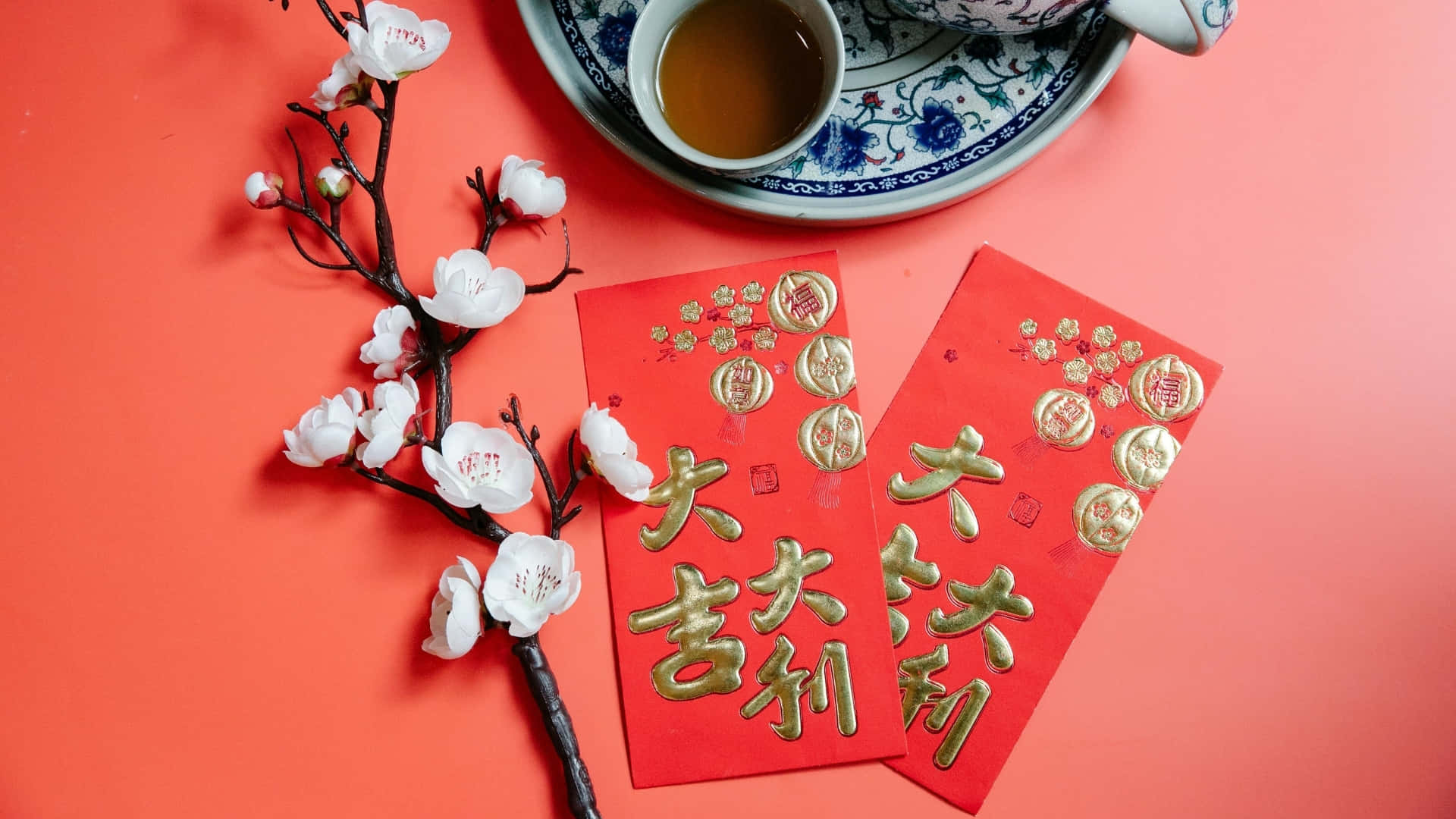  壁紙 — 紅色信封上印有金色中文字和白色櫻花，放在珊瑚色背景上的藍白茶杯旁邊。
