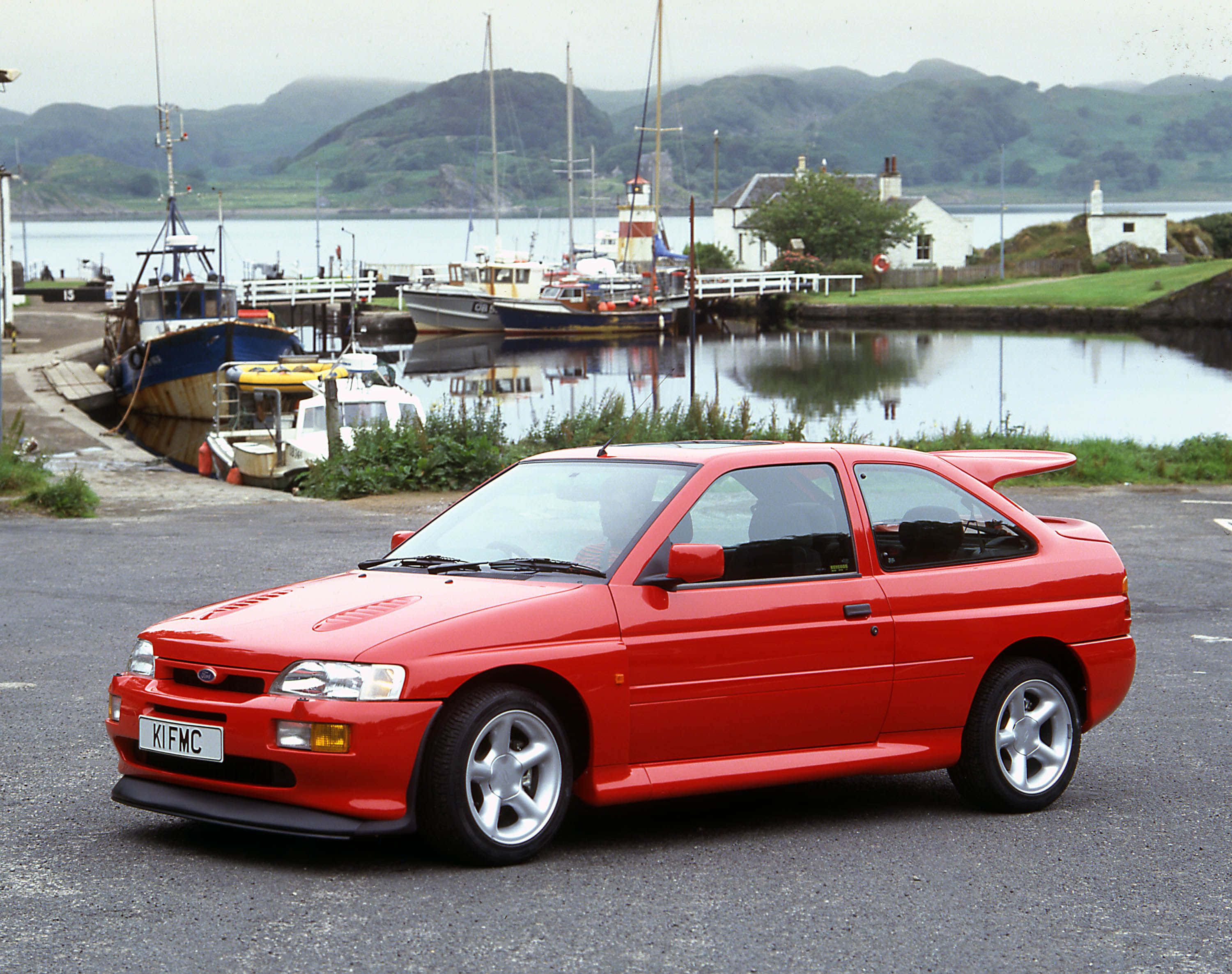 Ford Escort 桌布 壁紙 — 一架鮮紅色嘅福特 Escort RS Cosworth 泊喺瀝青路上，旁邊係有船同霧氣籠罩嘅綠色山丘嘅港口。