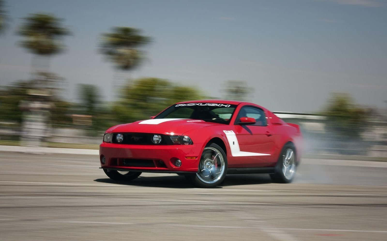 Ford Mustang Roush 桌布 壁紙 — 一輛紅色福特野馬在晴朗天空下飄移，留下煙霧，背景的棕櫚樹模糊不清。