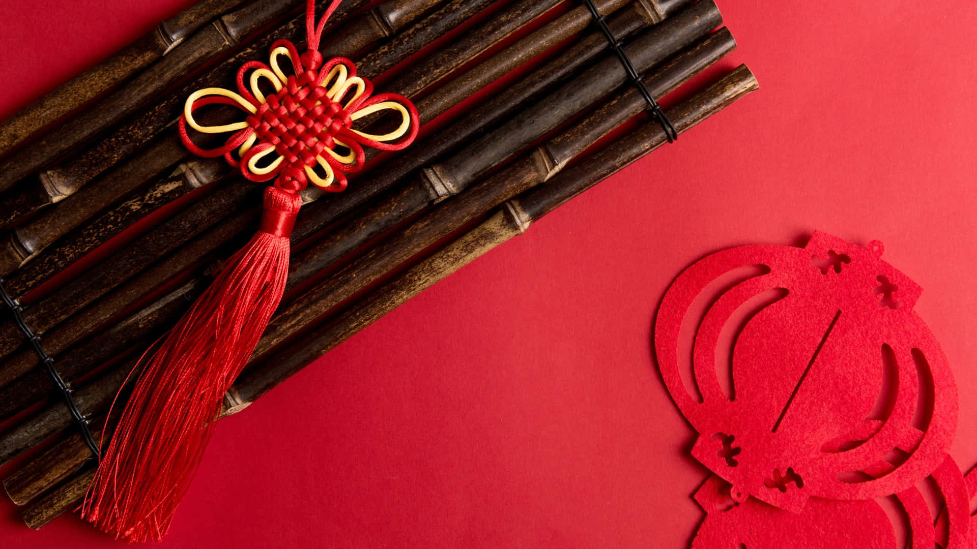  壁紙 — 一個紅色和金色的中國結飾品（帶流蘇）放在竹子上，背景是鮮豔的紅色，旁邊有一個紅色的紙燈籠剪紙。