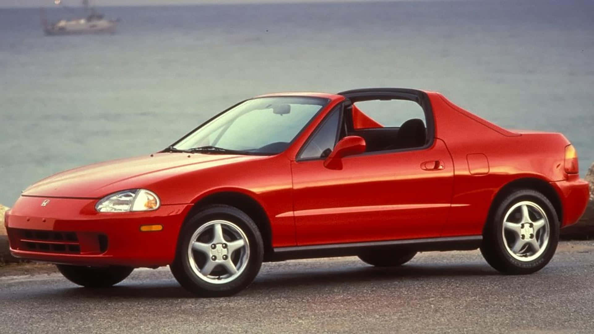 Honda Civic Del Sol 桌布 壁紙 — 一架鮮紅色嘅Honda Beat開篷車停泊喺海邊，遠處有帆船。