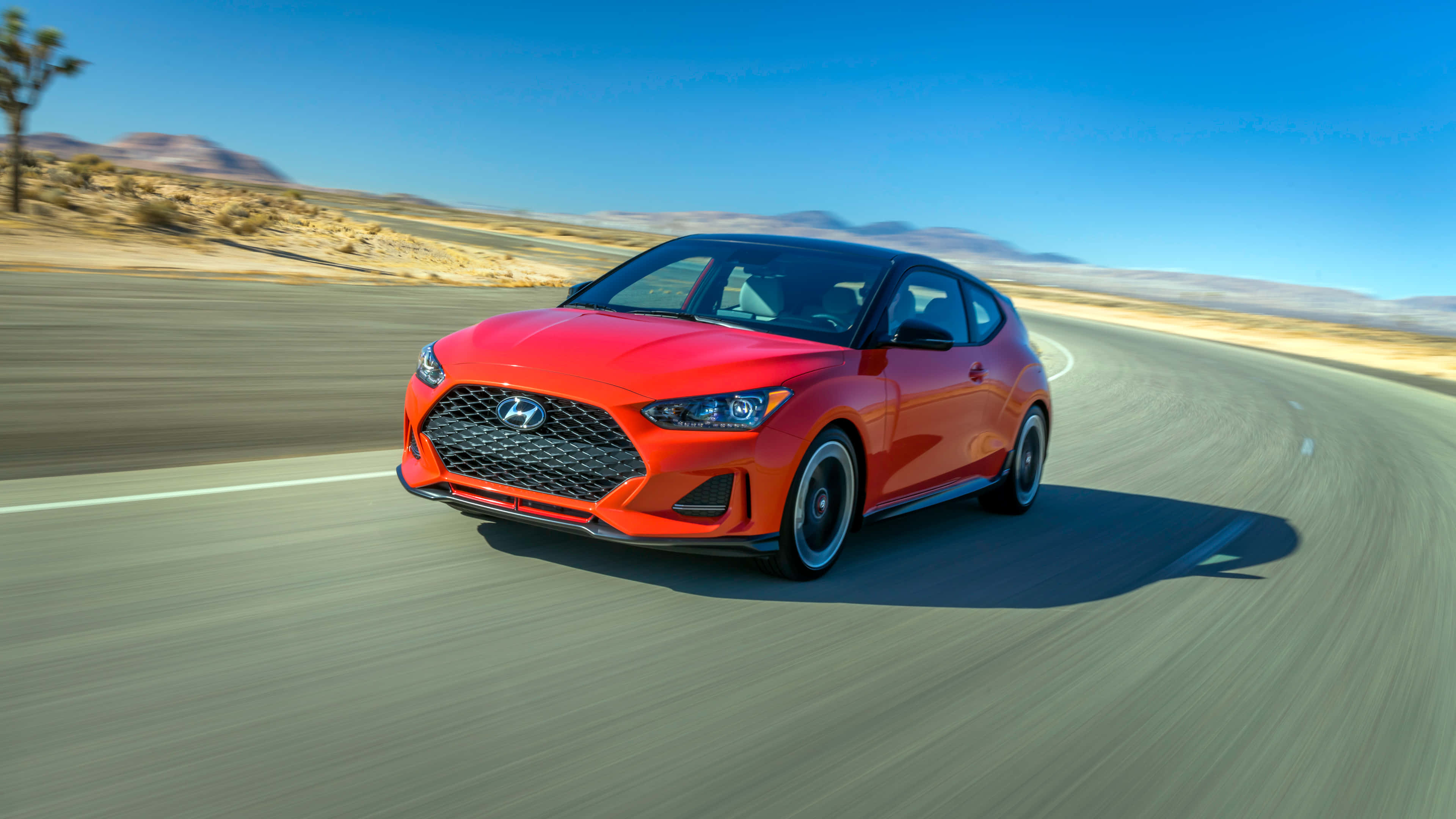 Hyundai Veloster 桌布 壁紙 — 一輛鮮紅色的現代Veloster在晴朗藍天和遠山背景下，沿沙漠高速公路飛馳。