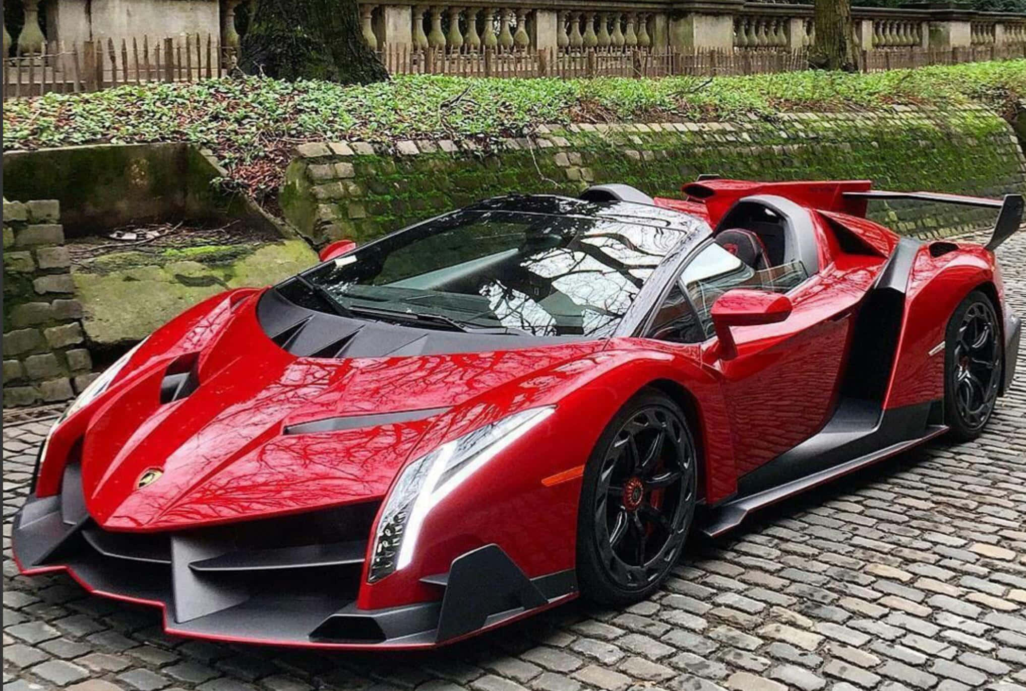 Lamborghini Veneno 桌布 壁紙 — 一架搶眼的紅色林寶堅尼 Veneno 開篷跑車停泊在鵝卵石街道上，旁邊是長滿青苔的石牆和綠植。
