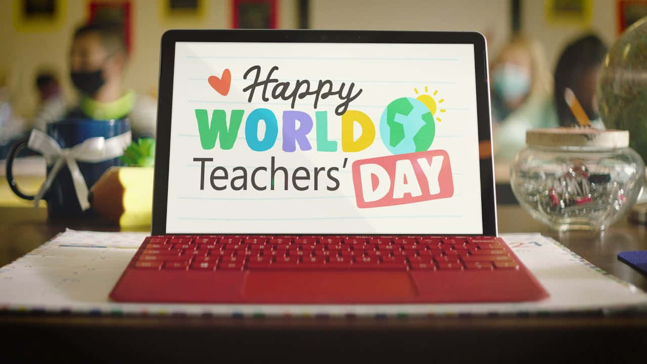 壁紙 — 一部紅色手提電腦在書桌上顯示色彩繽紛的「Happy World Teachers' Day」圖案，背景是模糊的課室。