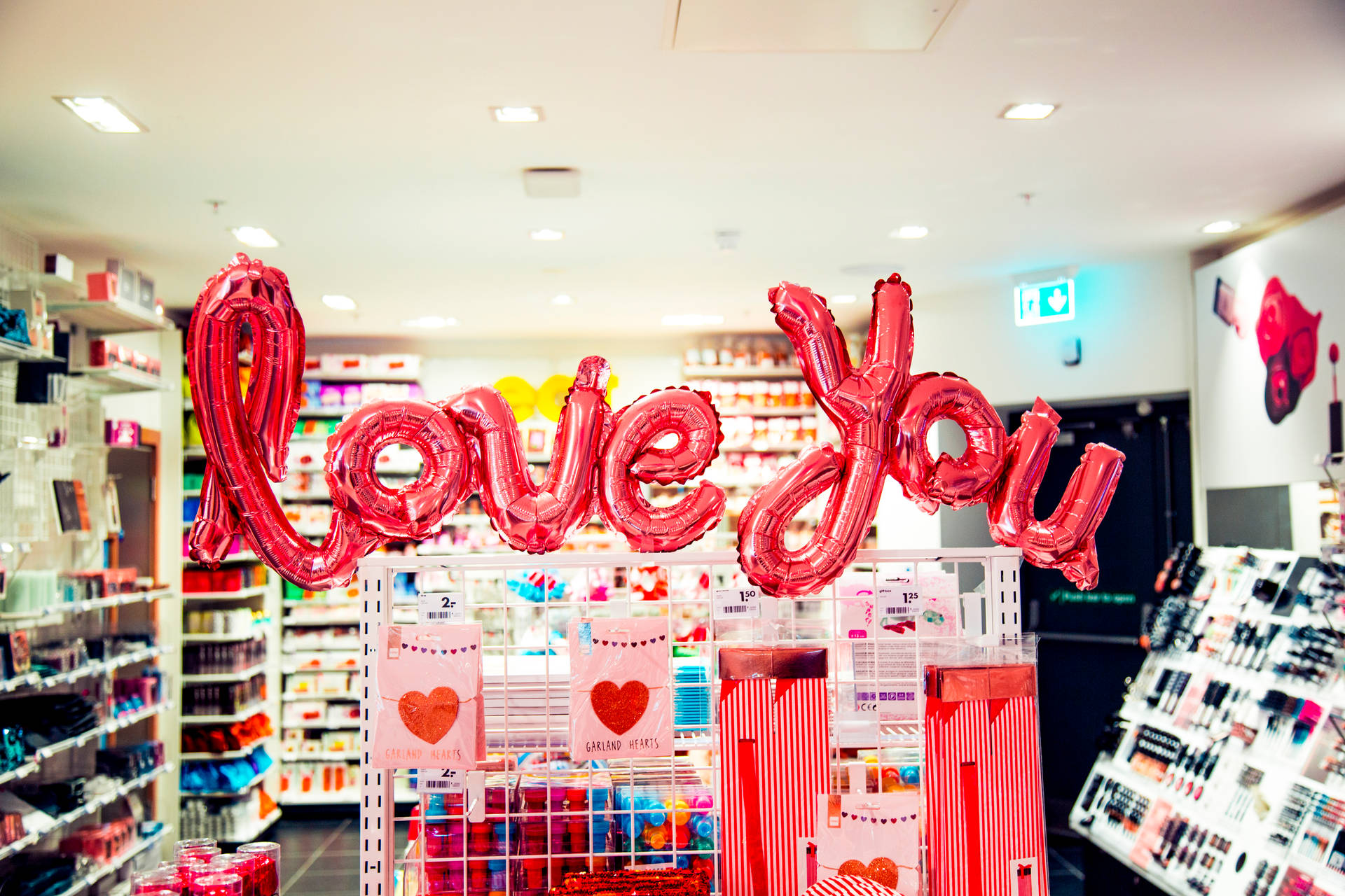 慶祝愛與浪漫的二月 壁紙 — 一個大型閃亮紅色 'Love You' 氣球漂浮在光線明亮的商店通道中的情人節展示上方。