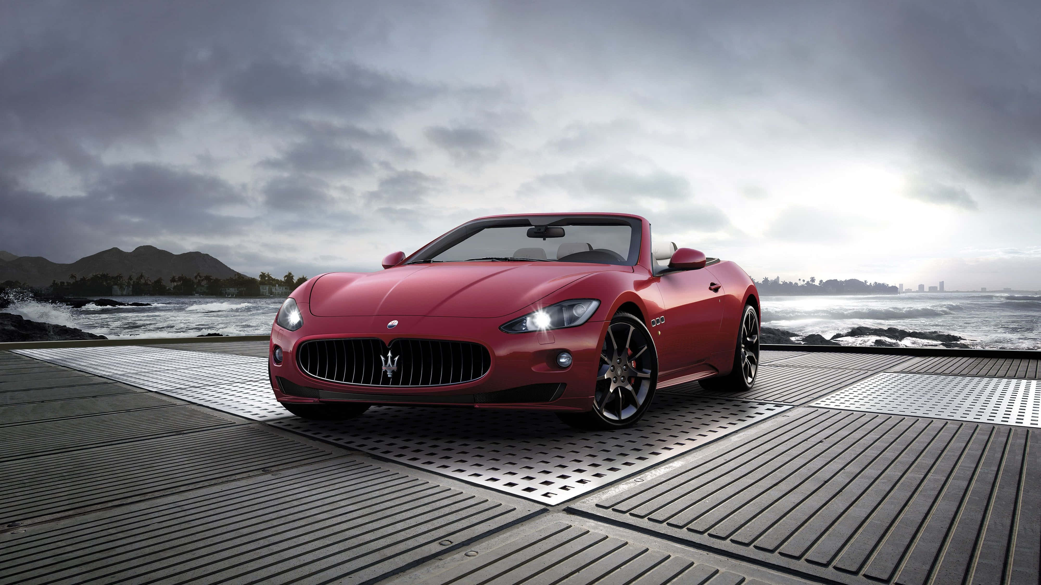 Red Maserati Parked 4K 壁紙 — 一架時尚的紅色開篷跑車停泊在金屬碼頭上，背後是洶湧的海浪和多雲的天空。