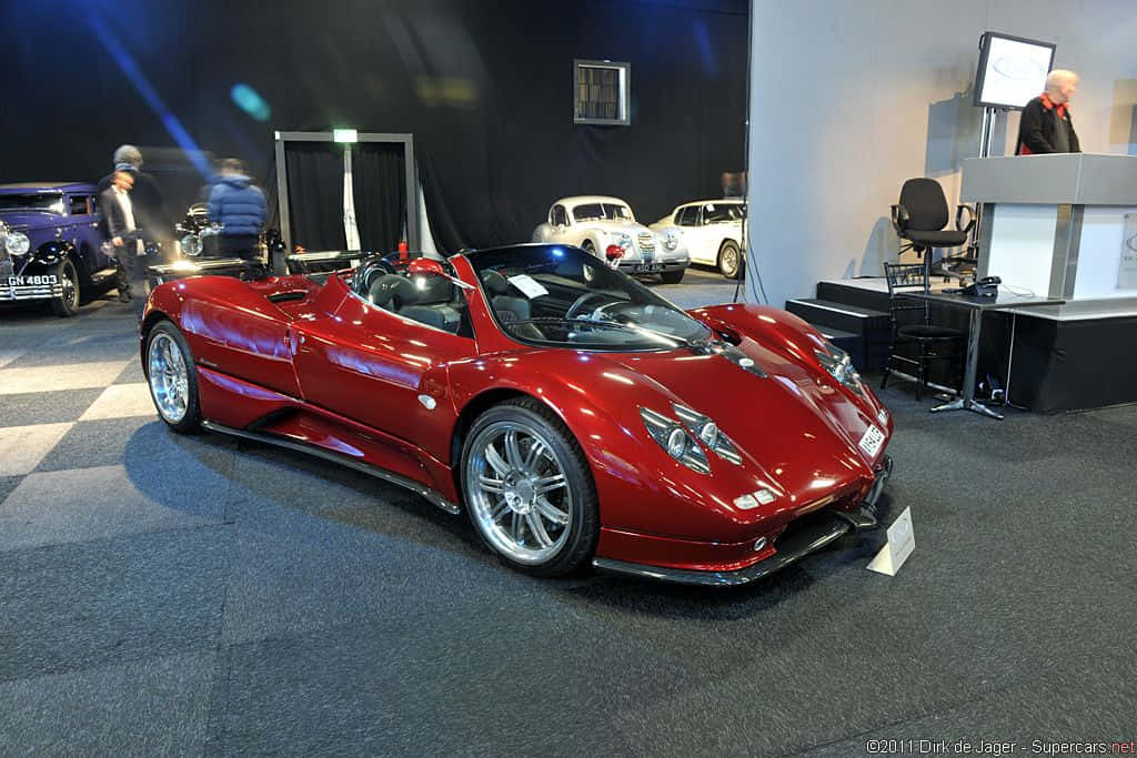Pagani Zonda C12 Roadster 桌布 壁紙 — 一輛光澤的紅色Pagani Zonda跑車在室內車展的聚光燈下閃耀，背景有其他經典車輛。