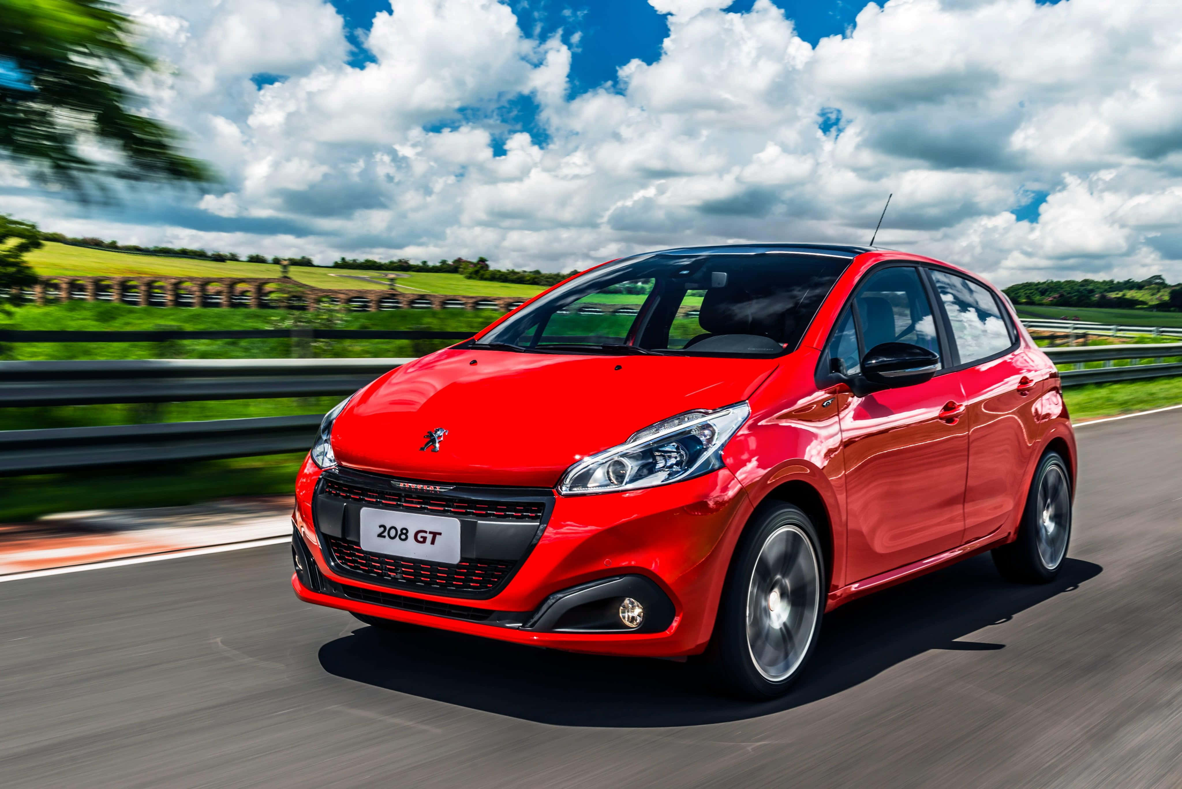 Peugeot 208 桌布 壁紙 — 一輛鮮紅色的標緻208 GT在蜿蜒的道路上疾馳，天空湛藍，白雲朵朵。