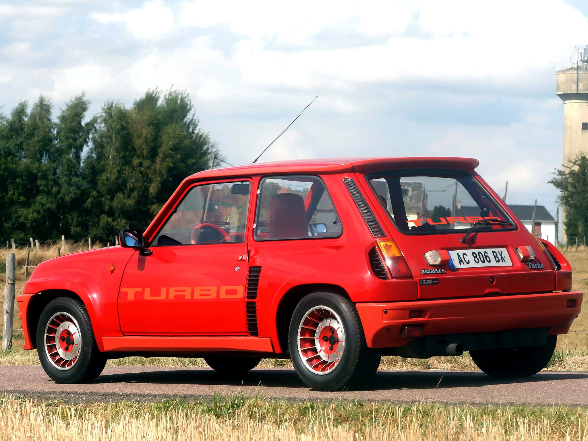 Renault 5 Turbo 桌布 壁紙 — 一輛鮮紅色的雷諾5 Turbo停泊在鄉村道路上，背景有金黃色的草和一個水塔，天空多雲。