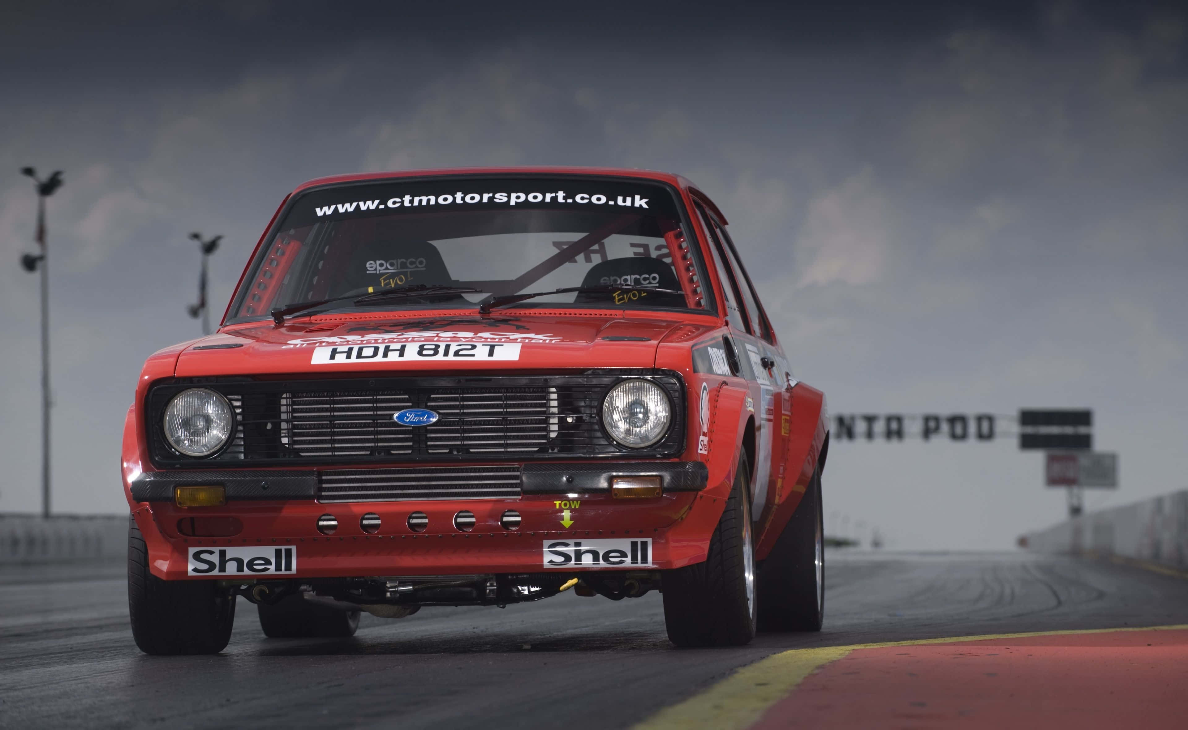 Ford Escort 桌布 壁紙 — 一輛紅色復古福特拉力賽車在暴風雨天空下的潮濕賽道上向鏡頭疾馳。