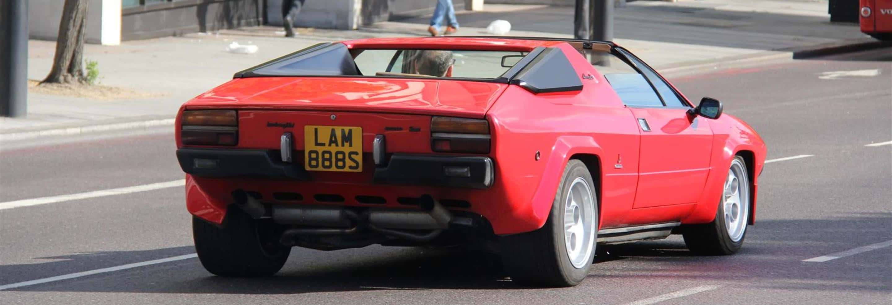 Lamborghini Urraco 桌布 壁紙 — 一輛鮮紅色的復古跑車在陽光明媚的城市街道上行駛，透過敞開的車頂可以看到司機。