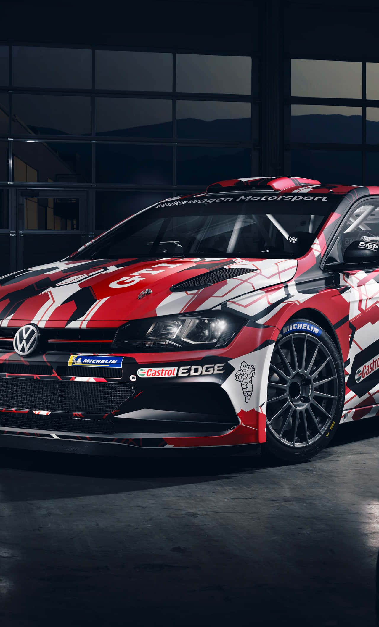 Volkswagen Polo 桌布 壁紙 — 一輛紅白黑色的福士汽車拉力賽車，帶有贊助商貼紙，停放在光線昏暗的車庫內，窗外可見山脈。