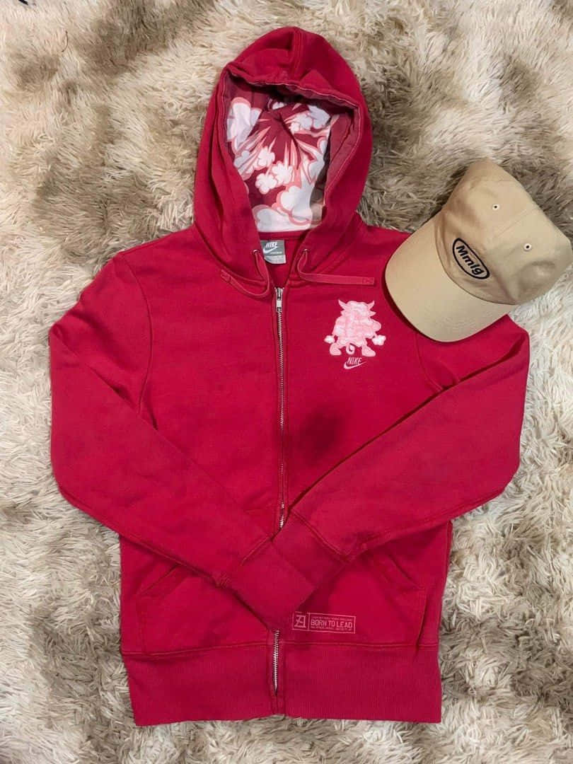 Red Hoodie 桌布 壁紙 — 一件紅色拉鍊連帽衛衣，帶有花卉內襯和粉紅色豬標誌，平放在柔軟的米色地毯上，旁邊有一頂米色帽子。