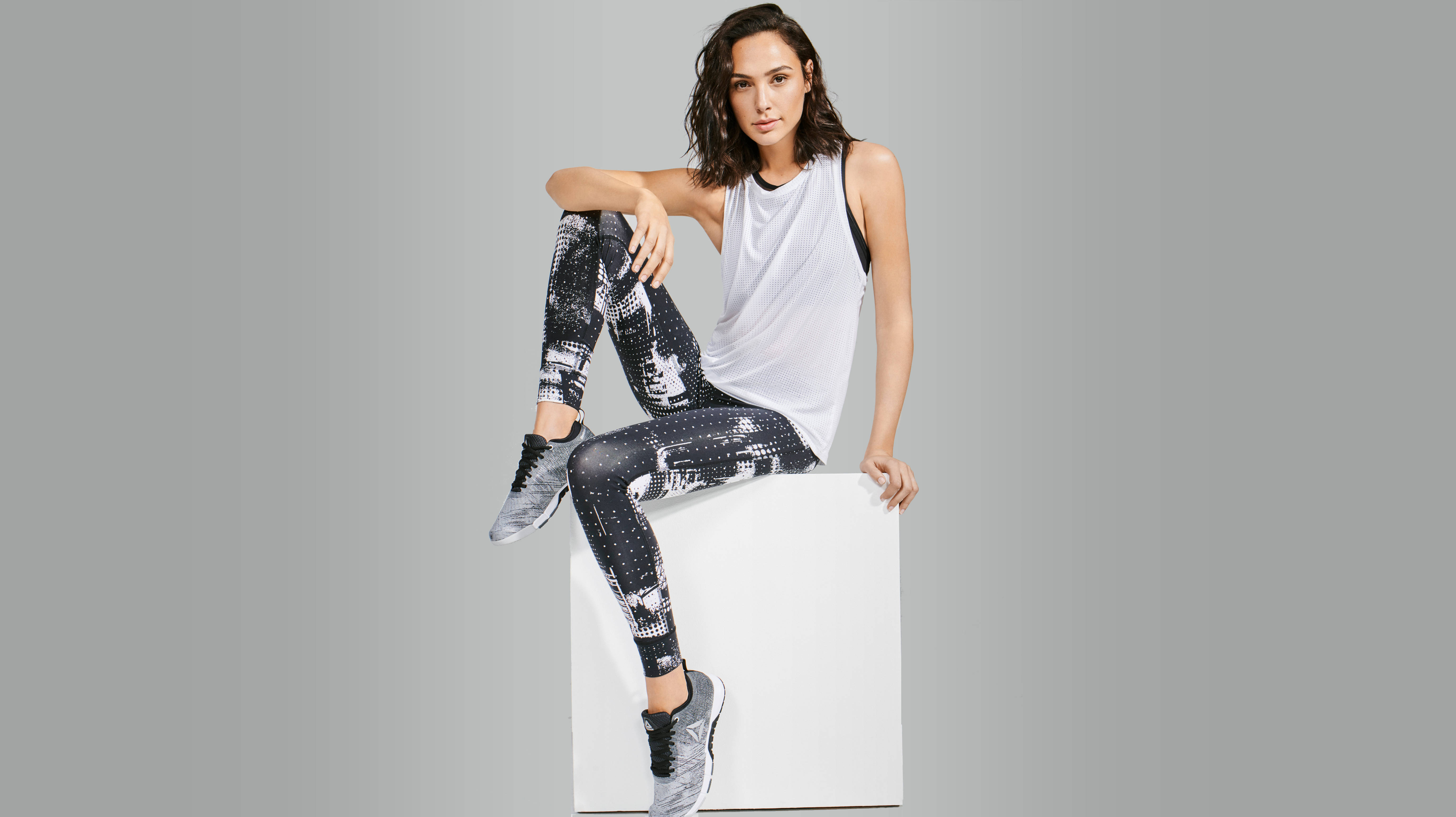 蓋兒·加朵為Reebok 2018 壁紙 — 一位穿著運動服的女性自信地坐在白色立方體上，背景為中性灰色。