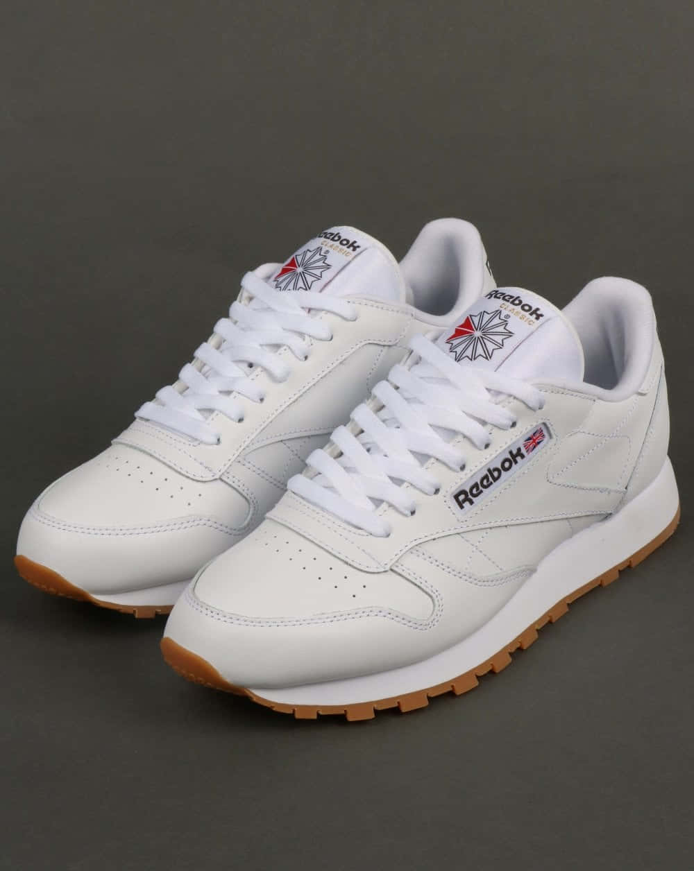穿上Reebok鞋履，展現最佳運動姿態 壁紙 — 一對純白Reebok Classic運動鞋配搭橡膠鞋底，整齊地斜放在深灰色工作室背景上。