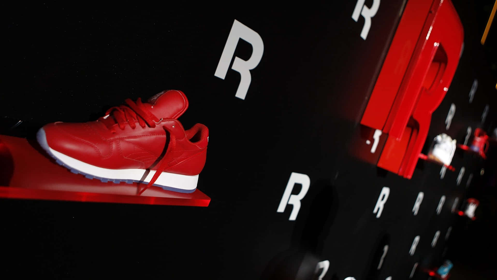以Reebok時尚動起來 壁紙 — 一雙大膽的紅色運動鞋在聚光燈下，在相配的紅色架子上，對著印有重複白色R標誌的黑色牆壁閃閃發光。