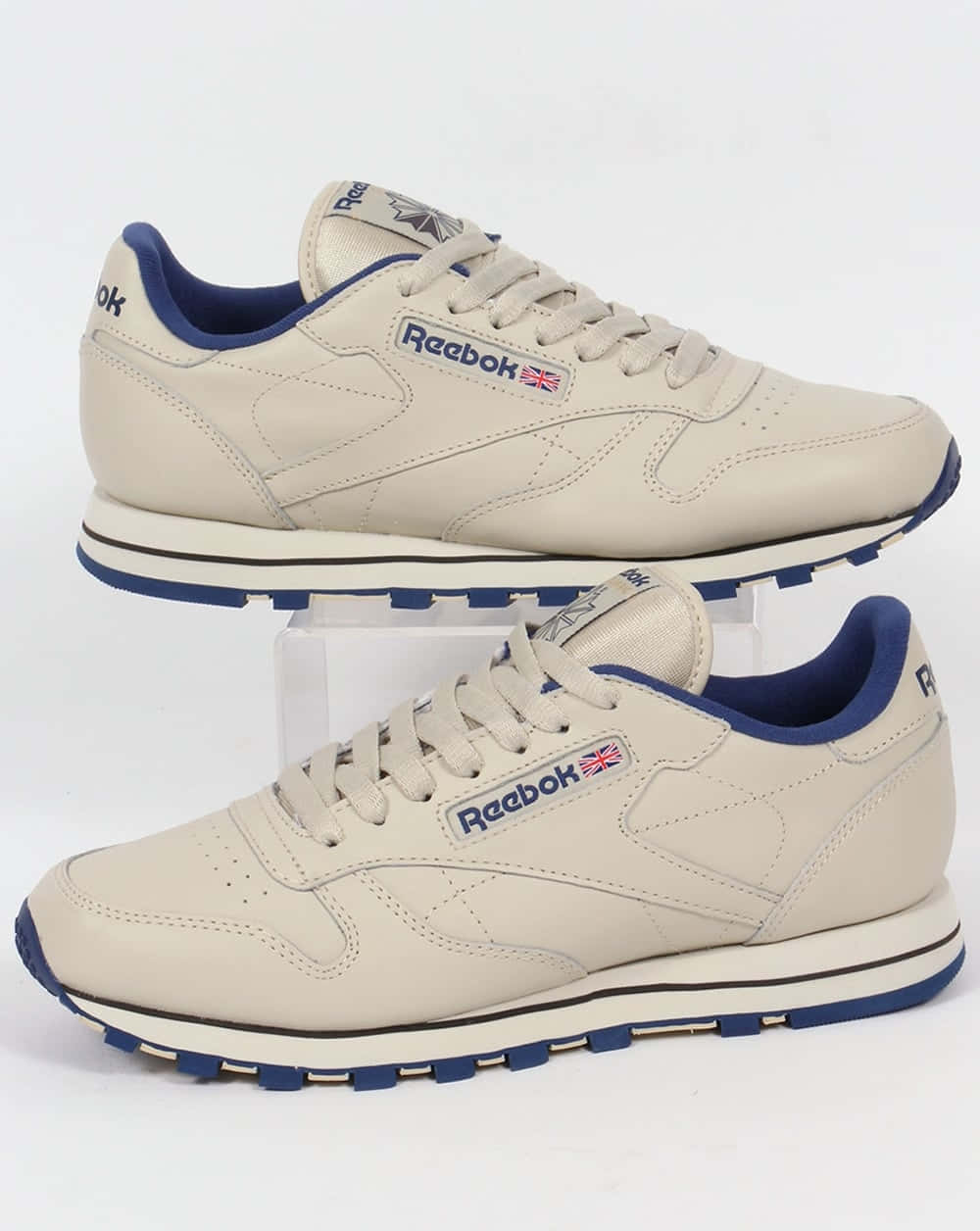 以Reebok展現你的個人風格 壁紙 — 兩雙米色 Reebok Classic 運動鞋，帶有藍色點綴，展示在白色方塊上，背景為純白色。