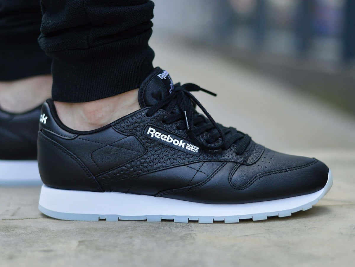 用Reebok把你的健身表現提升到新境界 壁紙 — 黑色Reebok Classic運動鞋白色鞋底在混凝土行人路上的特寫。