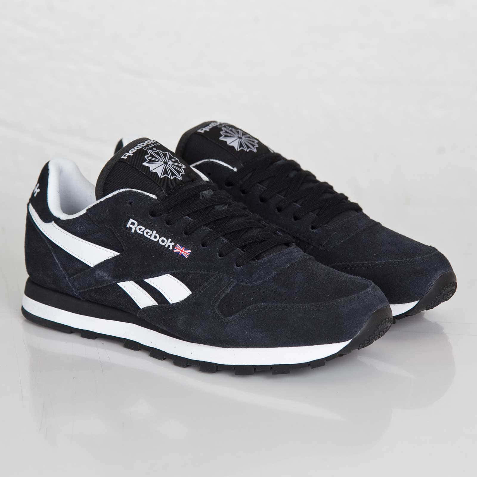 穿上Reebok，時刻保持活力四射！ 壁紙 — 一對黑色麂皮Reebok運動鞋，帶有白色條紋和英國國旗細節，放在反光的白色表面上。