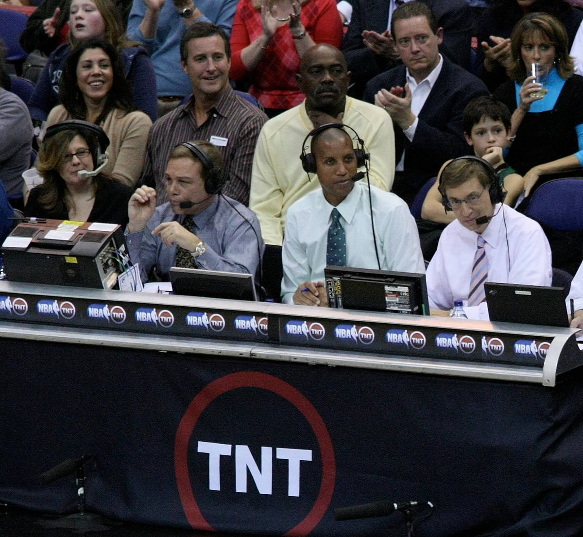 雷吉·米勒擔任場邊記者 壁紙 — 三位戴著耳機的NBA TV評論員坐在TNT品牌的廣播桌前，身後是熱鬧的人群。