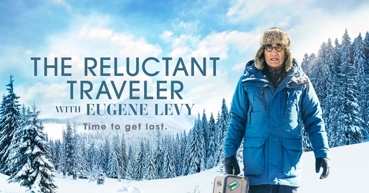 《Reluctant Traveler》電視劇橫幅海報 壁紙 — Eugene Levy 站在雪地森林中，身穿藍色冬季外套和毛帽，手提行李箱，背景是明亮的藍天白雲。