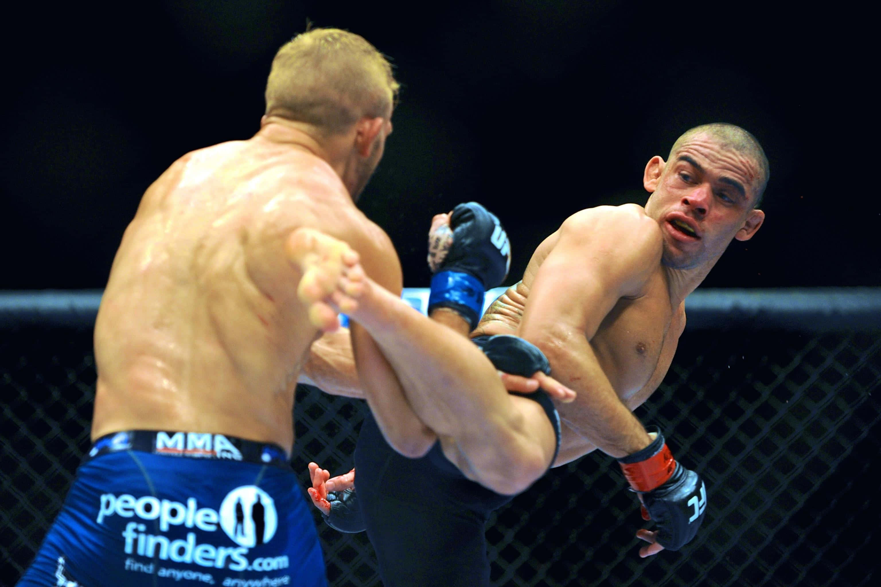 Renan Barão踢擊TJ Dillashaw 壁紙 — 兩名MMA拳手在籠中交鋒，一人使出強力踢擊，另一人則在汗流浹背的激烈時刻阻擋。