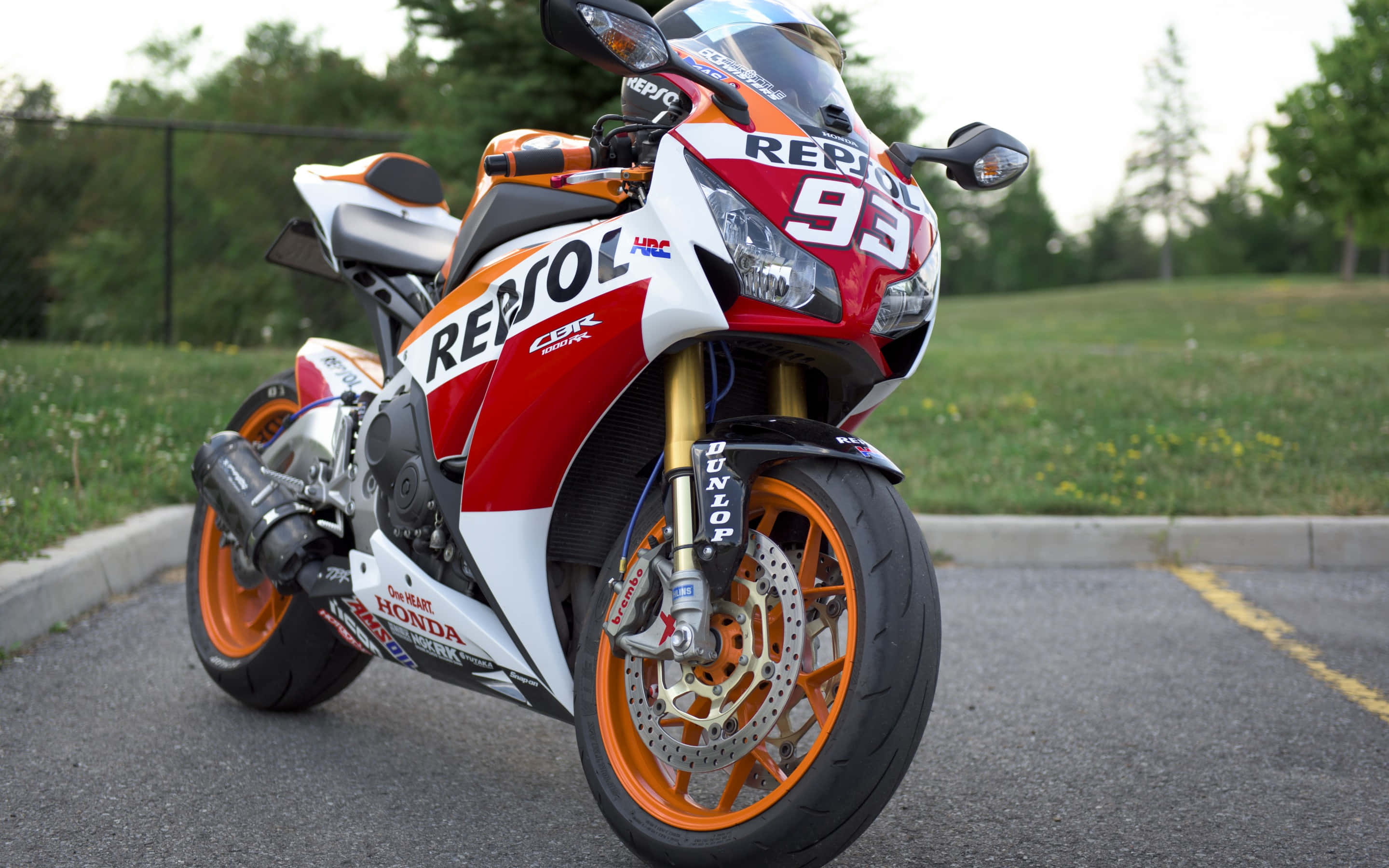 Repsol 超級摩托車圖片 壁紙 — 一架紅色、白色和橙色的Repsol Honda CBR賽車電單車停在瀝青路上，背景有綠草和樹木。
