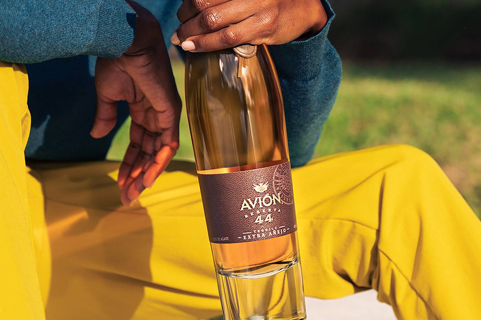 戶外穿著亮黃色褲子的人拿著一瓶 Avion 龍舌蘭酒。
