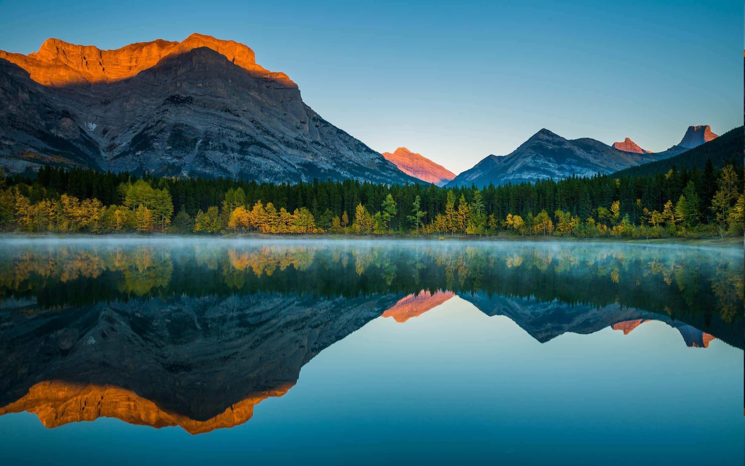 Fall Lake 桌布 壁紙 — 日出將山峰沐浴在金色光芒中，完美地倒映在被秋樹環繞的霧氣瀰漫的湖面上。