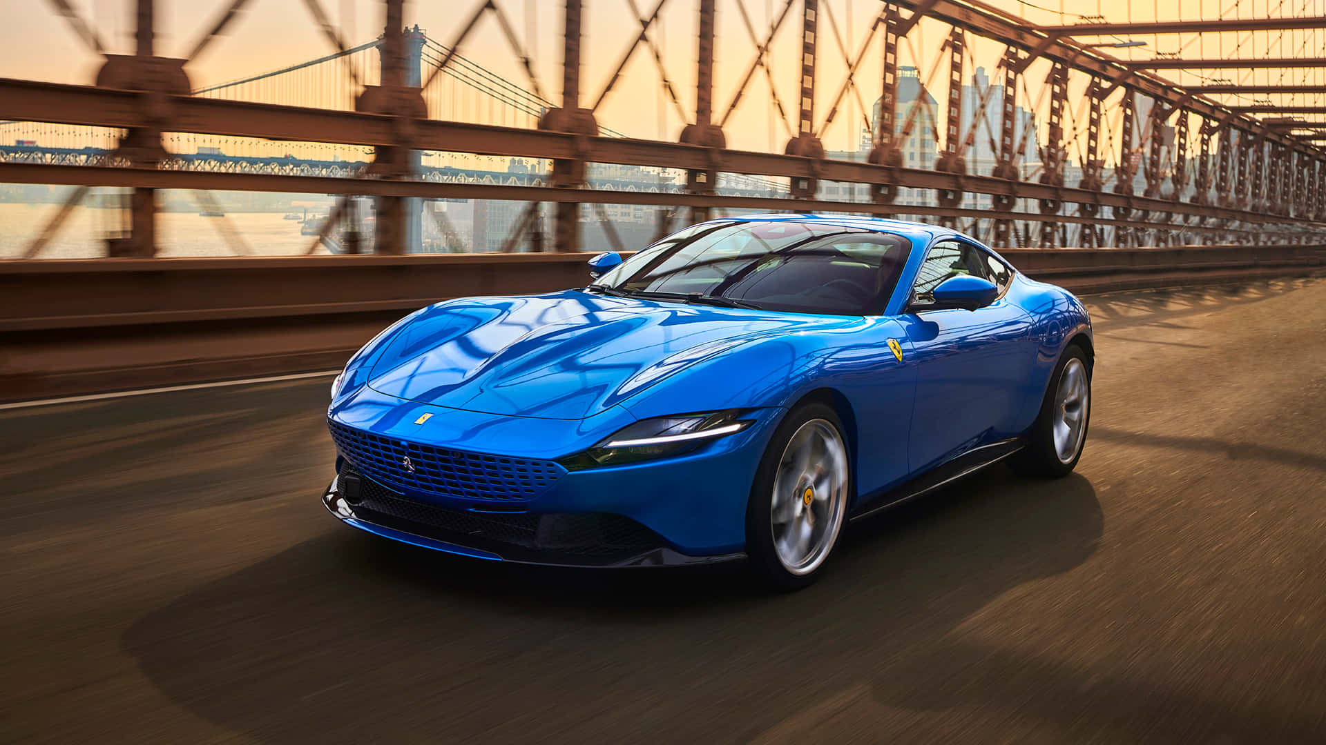 Ferrari Roma 桌布 壁紙 — 一輛充滿活力的藍色法拉利跑車在金色光線下，帶着動態模糊效果飛馳過一座鋼橋。