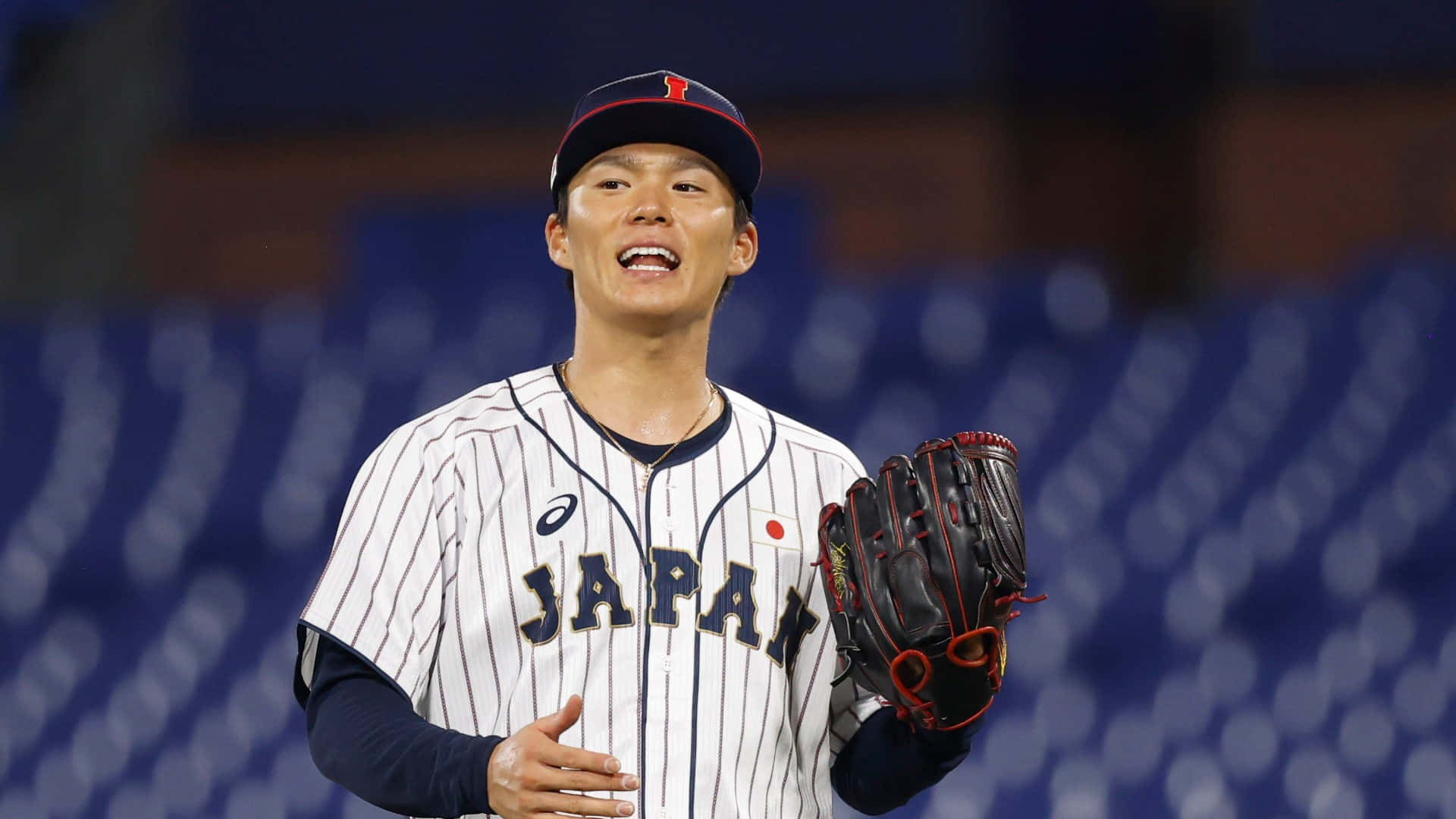  壁紙 — 一位身穿細條紋球衣的日本棒球選手，在模糊的球場背景下，手持手套露出燦爛笑容。
