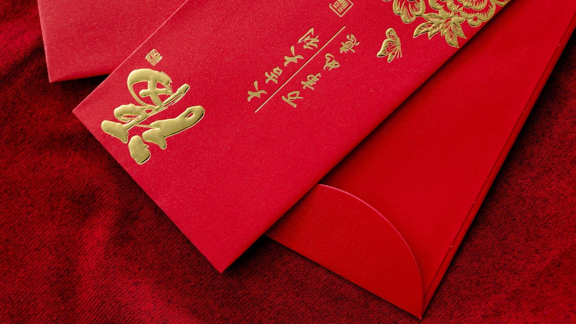 壁紙 — 豐富的紅色信封，帶有金色中國書法和花卉浮雕，放置在帶紋理的紅色織物表面上。