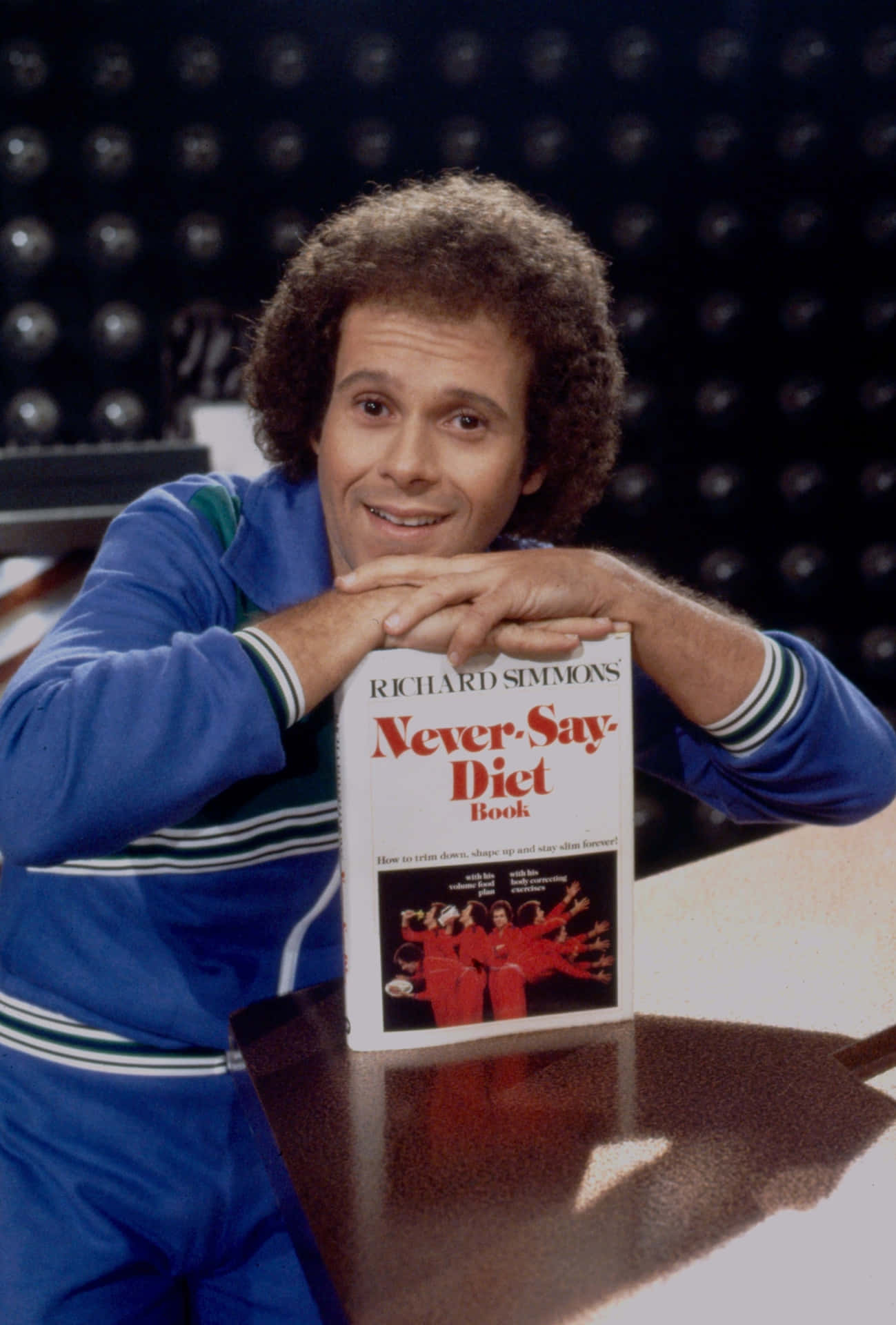  壁紙 — Richard Simmons 喺一個深色圓點背景嘅工作室入面，燦爛咁笑住攞住佢本《Never-Say-Diet》書。
