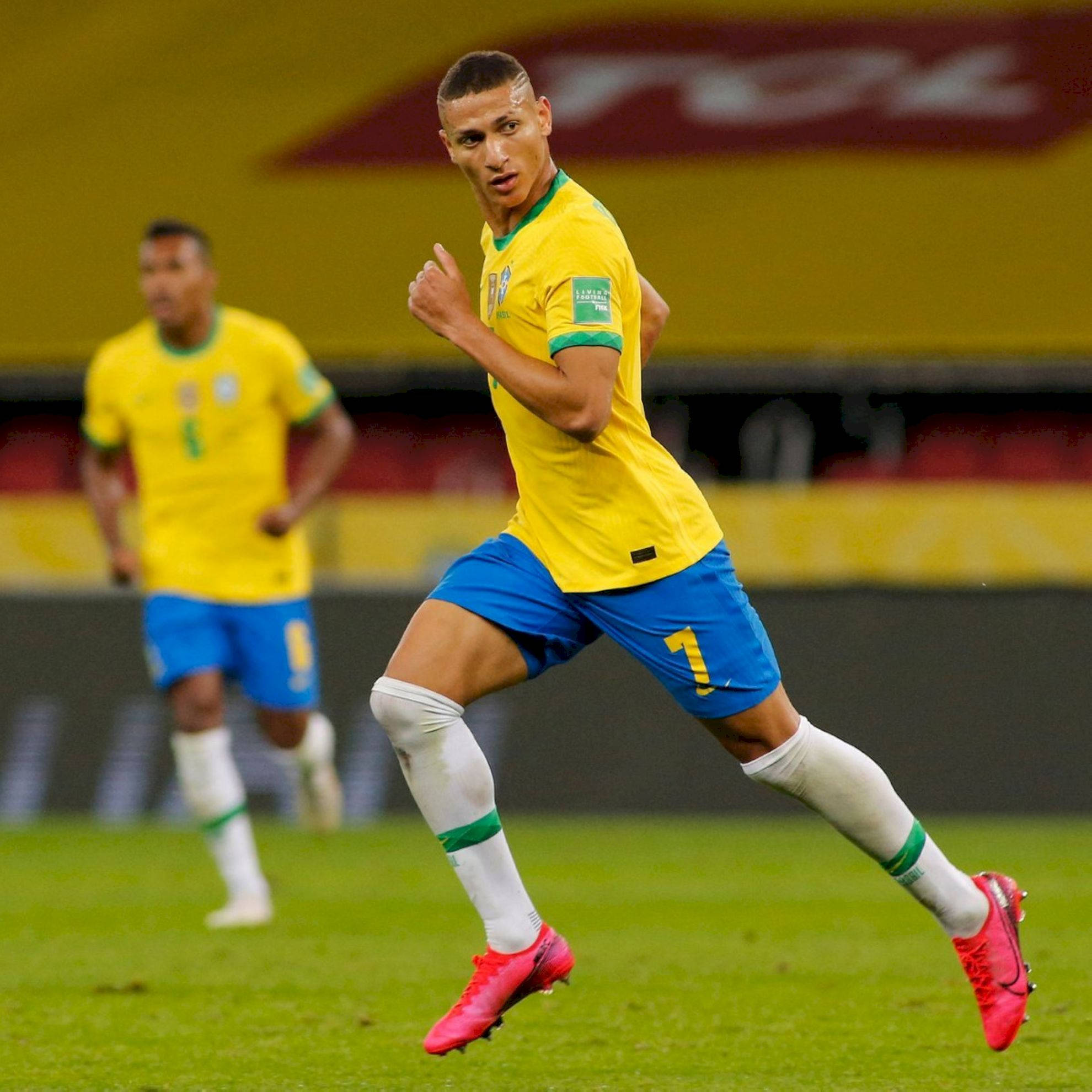 Richarlison De Andrade 奔跑 壁紙 — 一名身穿黃藍球衣的巴西足球員在夜間比賽中，專注地衝過綠色球場。