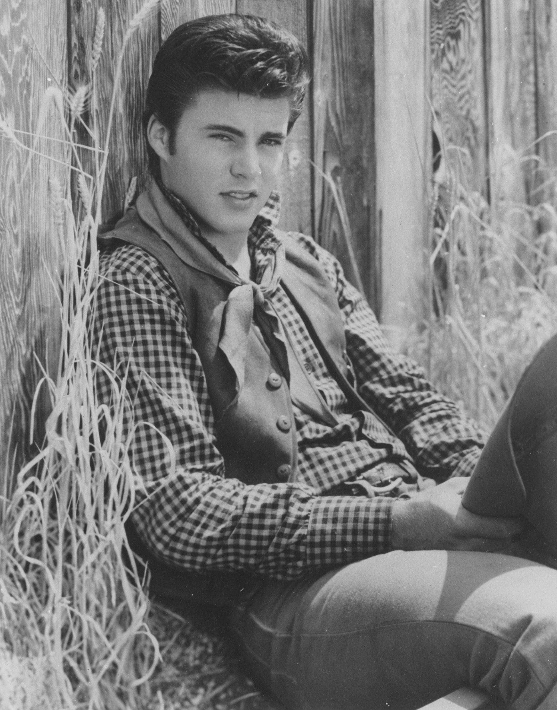 Rick Nelson《Rio Bravo 1959》黑白劇照 壁紙 — 一位身穿格仔恤衫和背心的年輕男子，隨意地靠著木柵欄坐著，周圍是高高的草叢，一幅復古黑白肖像。