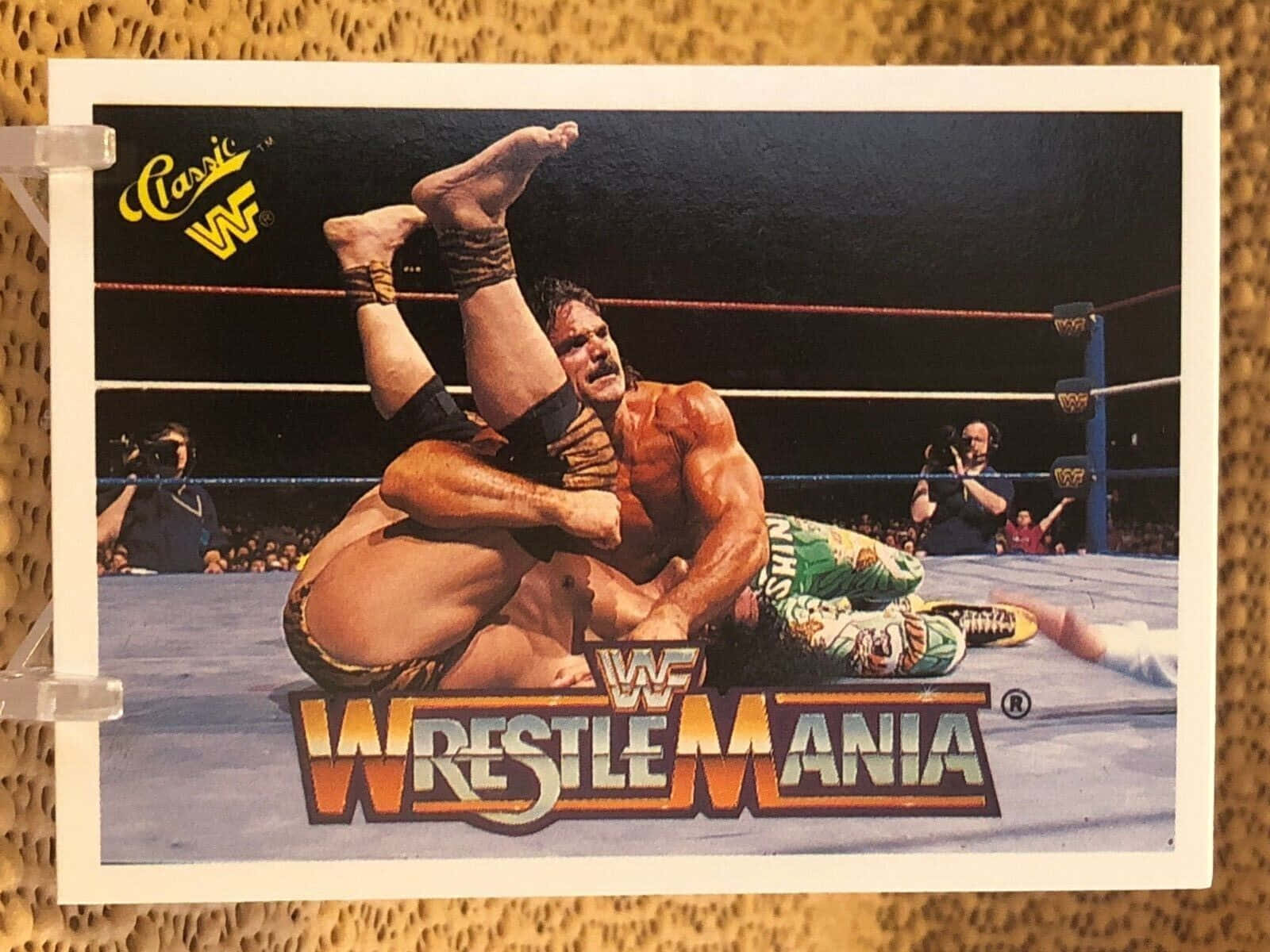 Rick Rude WWE WrestleMania 復古照片卡 壁紙 — 一位肌肉發達的摔角手在WrestleMania活動期間，在燈光明亮的摔角場內對對手執行腿部鎖定。