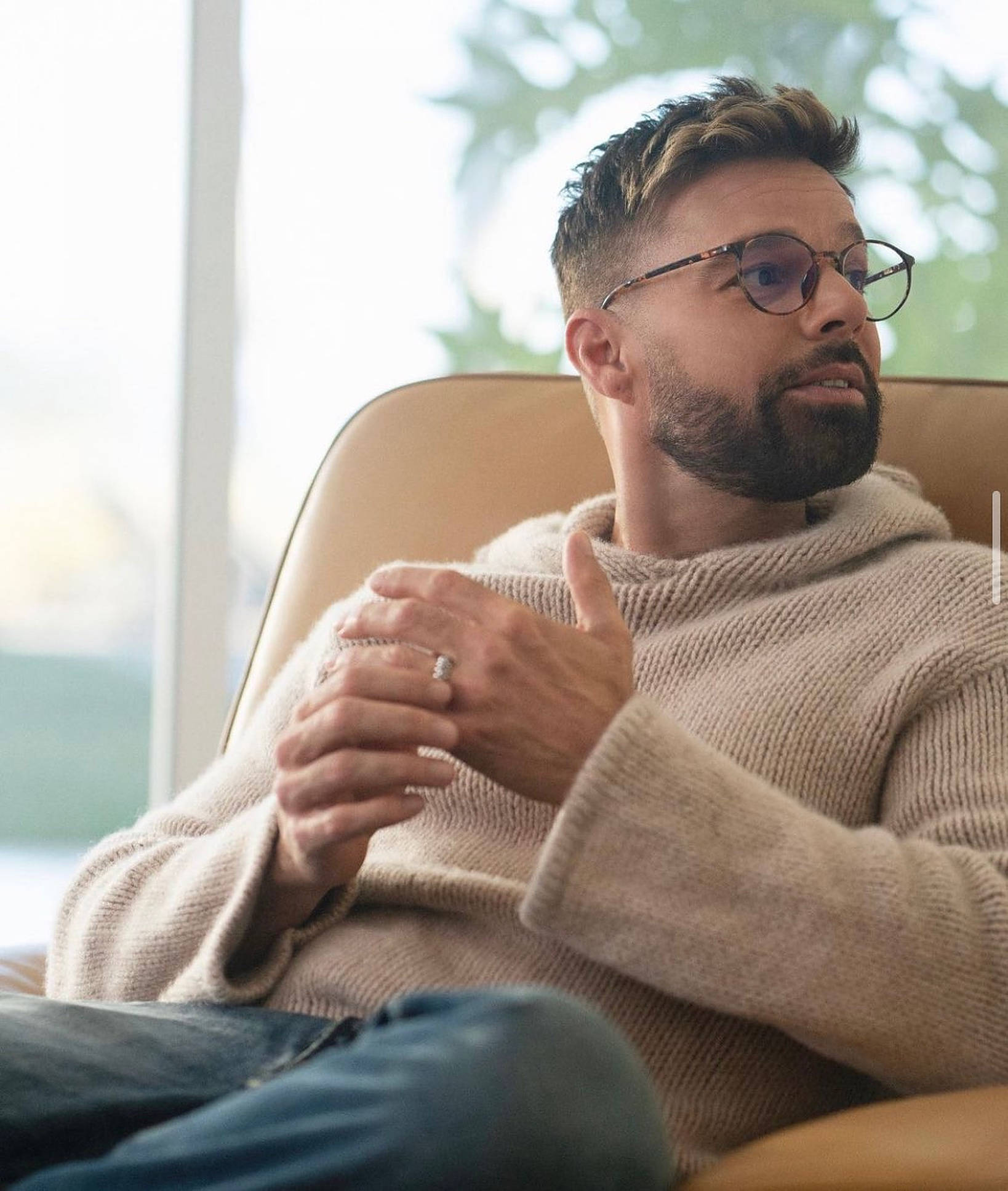 Ricky Martin 戴眼鏡 壁紙 — 一位戴眼鏡留鬍鬚的男人放鬆地坐在米色扶手椅上，穿著舒適的米色毛衣。