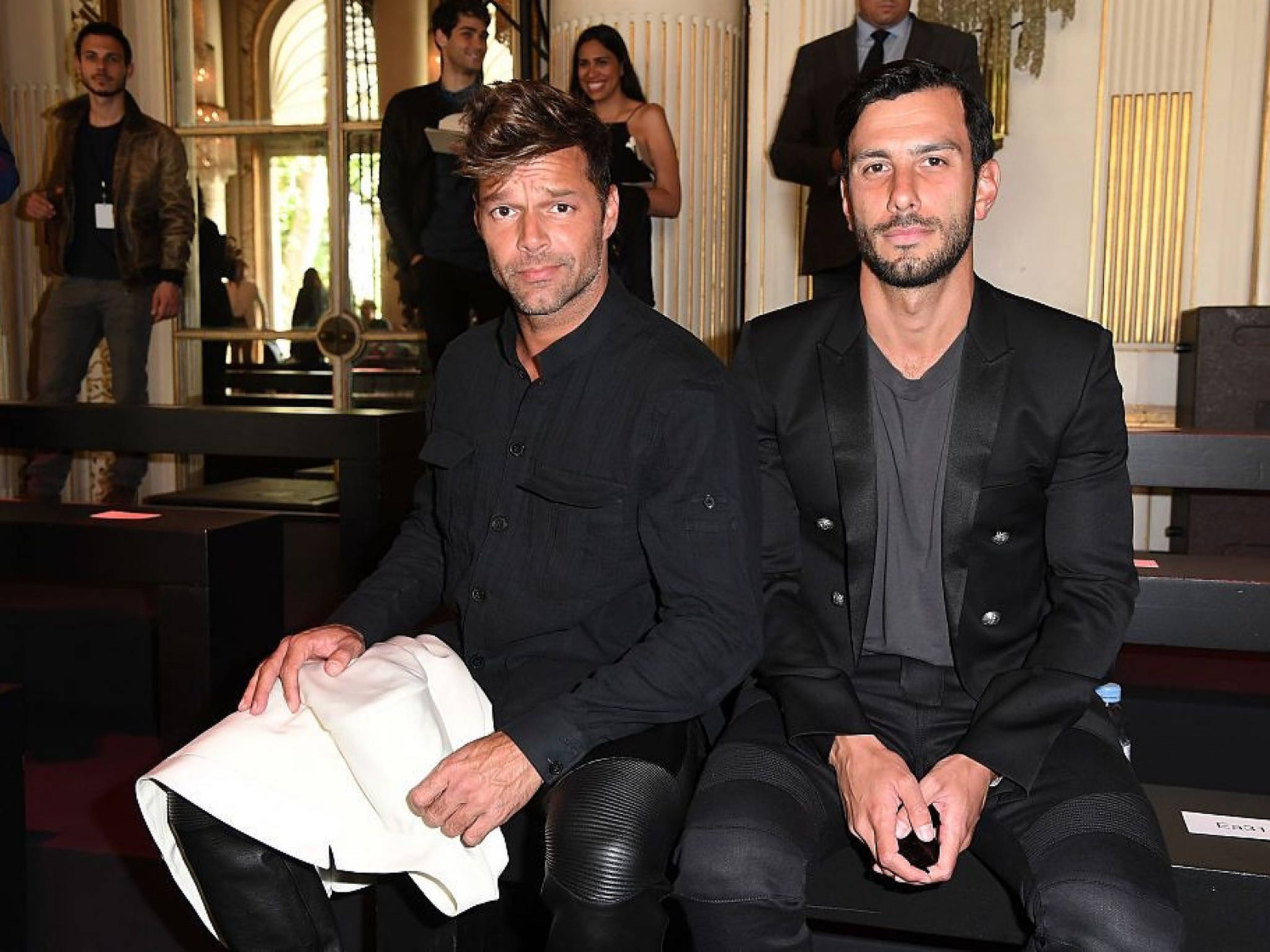 Ricky Martin 與 Jwan Yosef 壁紙 — 兩名身穿黑色西裝的男子在一個時尚活動中專注地坐著，背景有華麗的金色建築和其他與會者。
