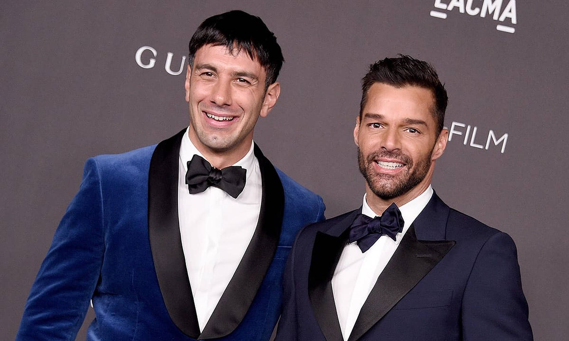 Ricky Martin 西裝領帶 壁紙 — 兩位微笑身穿燕尾服的男士在華麗的紅地毯活動中自信擺姿勢，背景帶有品牌標誌。