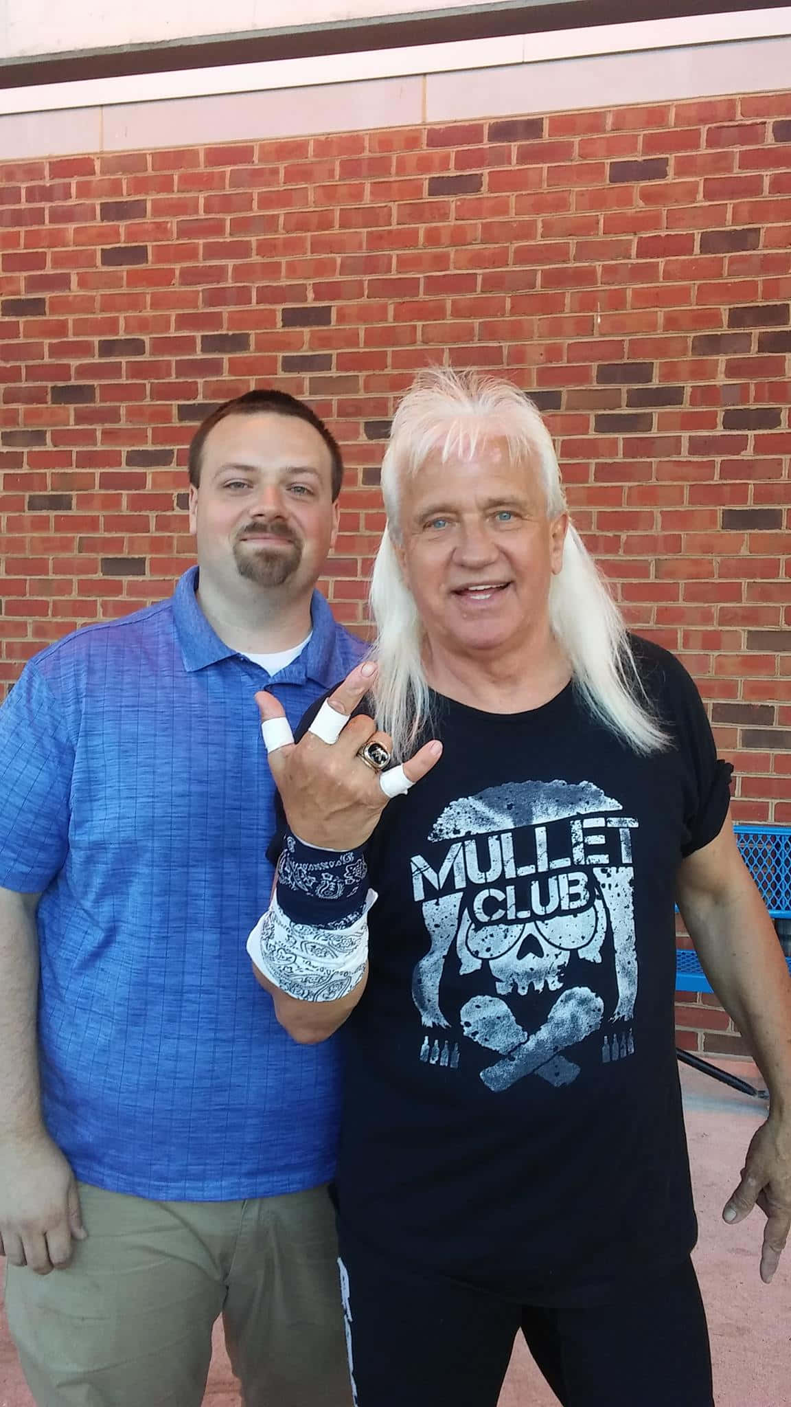 Ricky Morton 與粉絲合影 壁紙 — 兩個男人喺紅磚牆前面擺甫士，其中一個着住 mullet club T恤，做緊搖滾手勢。