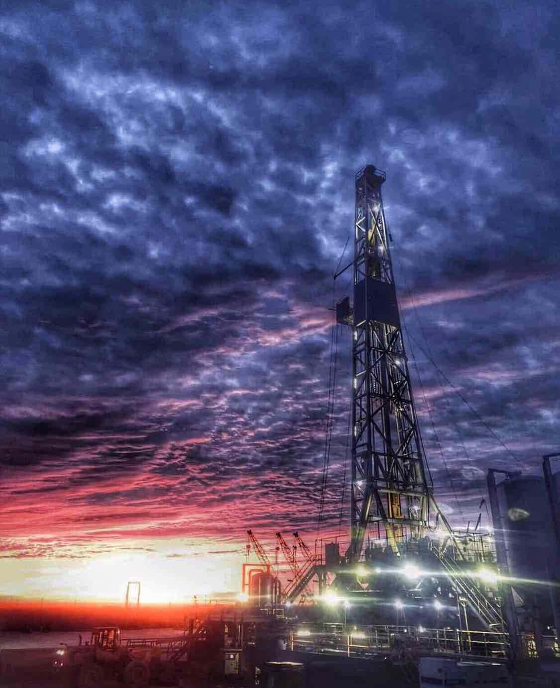 全球油 rigs 與氣候節約 壁紙 — 一座高聳的石油鑽井平台在戲劇性的暮色天空中亮起，天空佈滿火橙色和深紫色的雲彩。