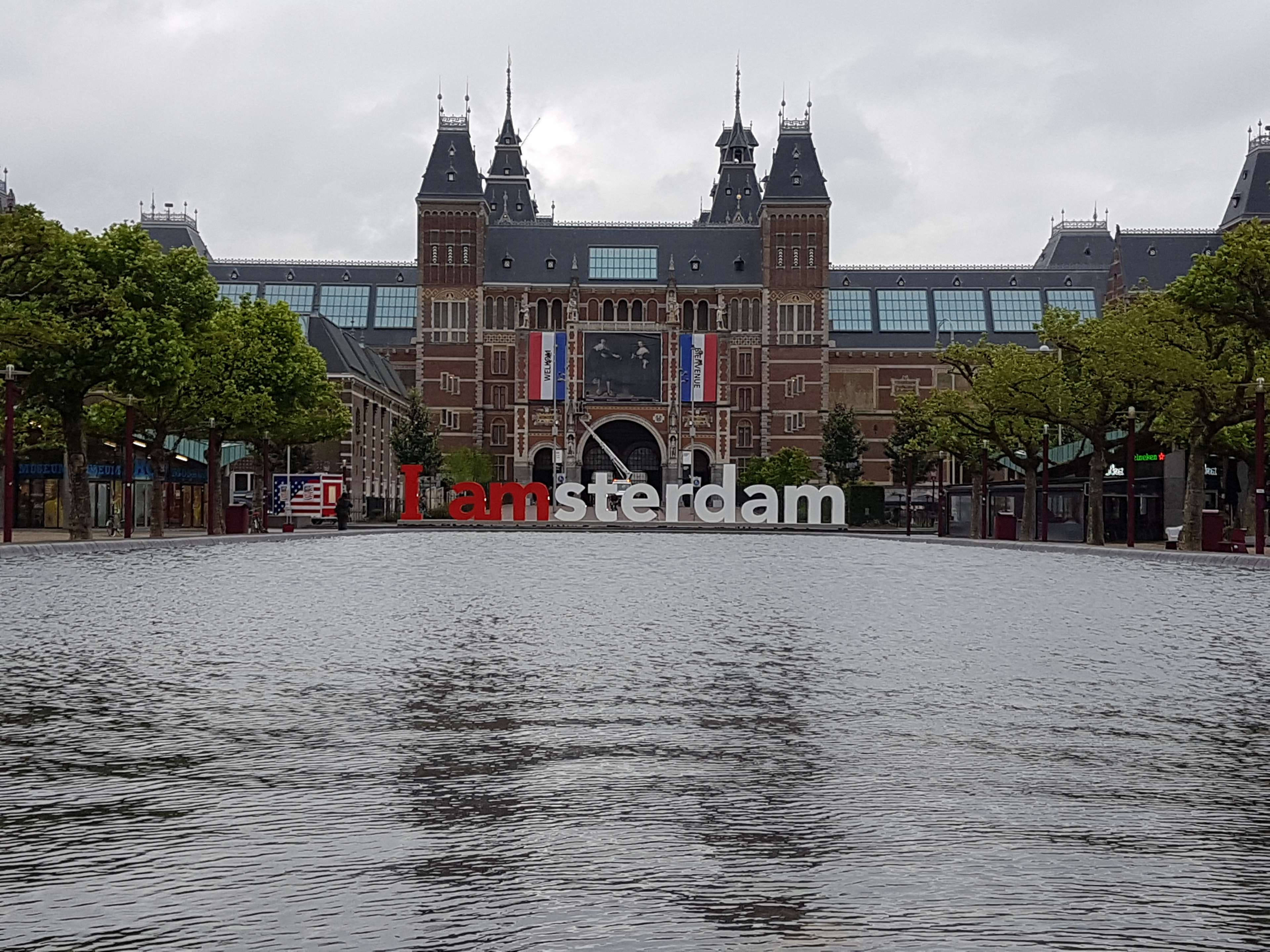 阿姆斯特丹 rijksmuseum 廣角陰天攝影 壁紙 — 一座宏偉的歷史阿姆斯特丹建築矗立在反射水景後面，前方有一個大型「I amsterdam」標誌。