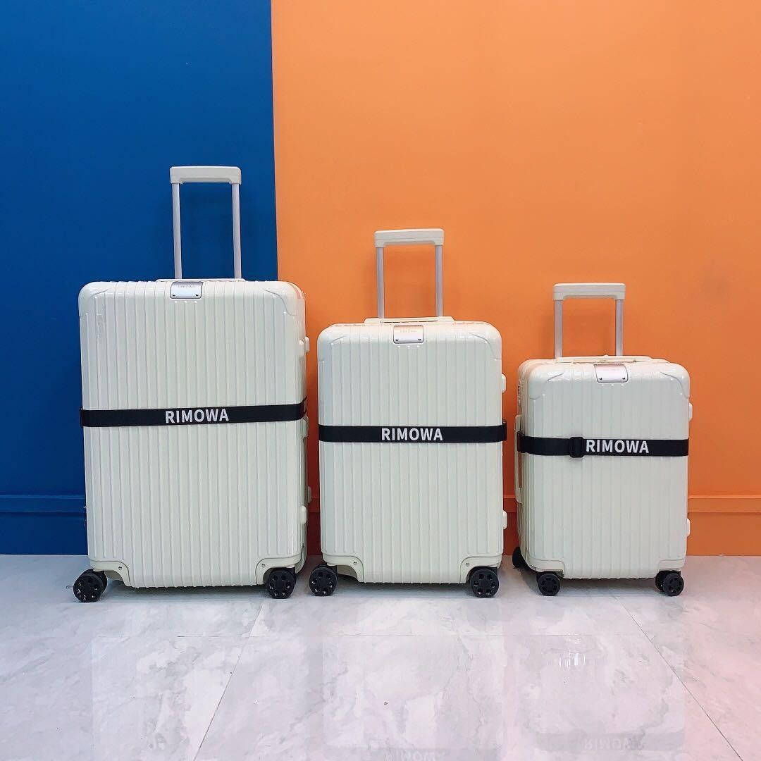 三件白色Rimowa行李箱 壁紙 — 三個帶有黑色帶子的奶油色Rimowa行李箱在大理石地板上排成一排，對著大膽的藍色和橙色牆壁。