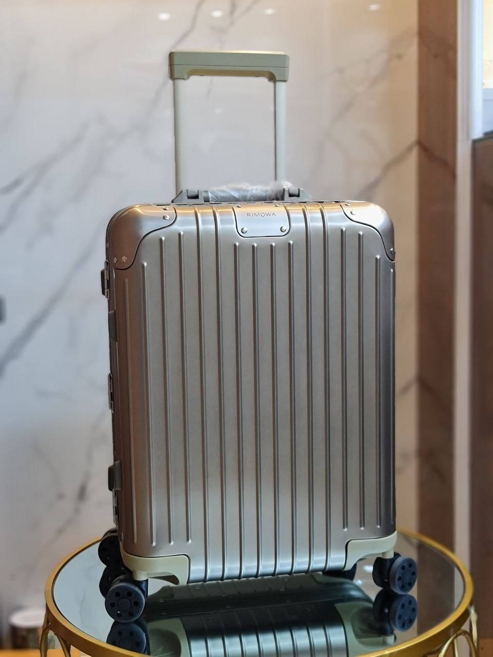 Rimowa 淡黃晶石機艙行李箱 壁紙 — 一個帶有垂直凹槽的時尚銀色Rimowa行李箱，直立放在大理石牆前的玻璃桌上。