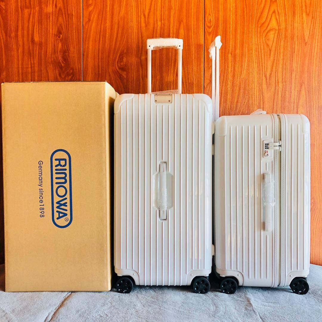 白色Rimowa行李箱 壁紙 — 兩個白色坑紋Rimowa行李箱站在其品牌紙箱旁邊，背景係溫暖嘅木牆。