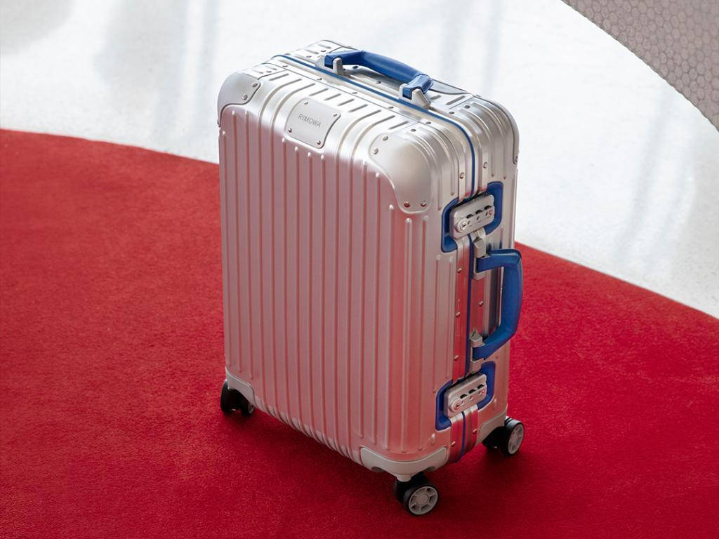 Rimowa Original Twist系列行李箱 壁紙 — 一個時尚的銀色手提箱，帶藍色手柄，放在柔軟的紅色地毯上，背景是白色垂簾。