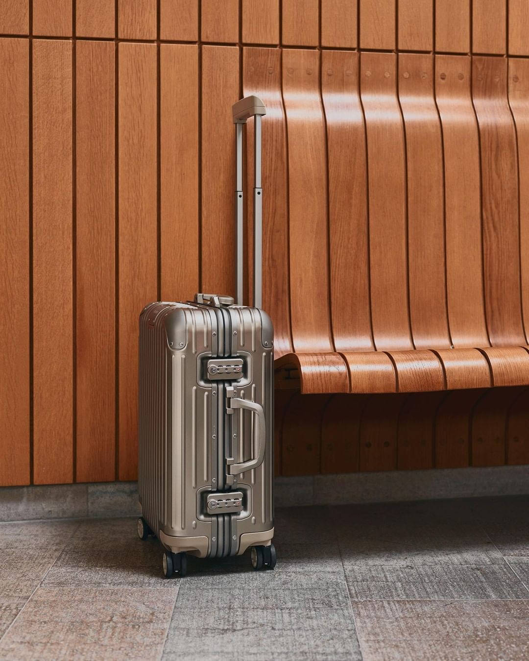 Rimowa原版行李箱 壁紙 — 一個時尚的銀色滾輪行李箱直立於現代室內環境中彎曲的木凳旁。
