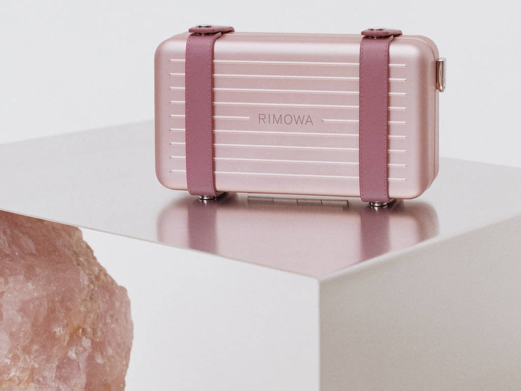 玫瑰金色Rimowa行李箱配紫紅色帶子，白色底座，粉紅色水晶旁。