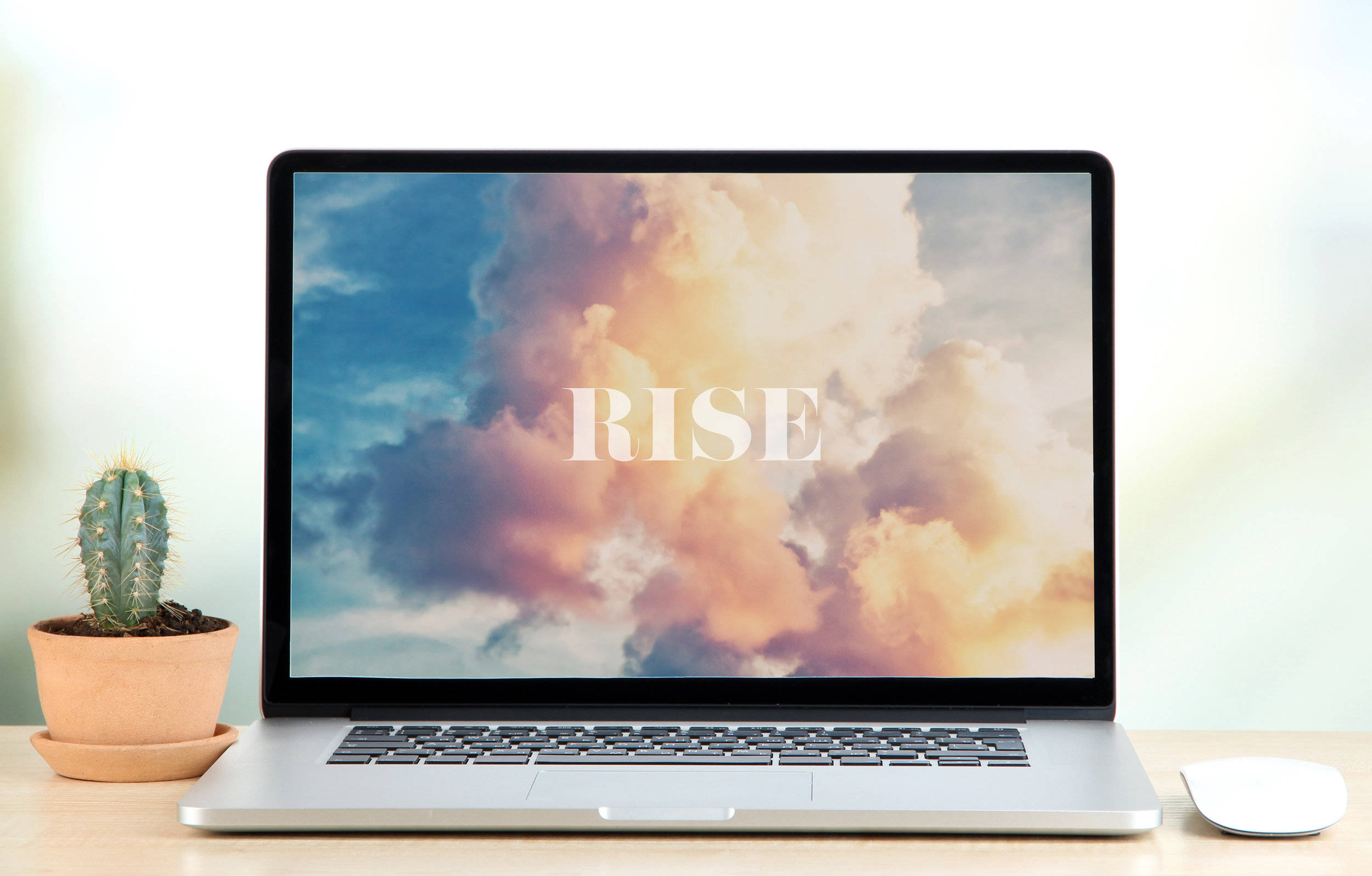 「Rise」字樣搭配雲朵背景的辦公室筆記本電腦壁紙 壁紙 — 一部時尚的筆記型電腦在充滿活力的日落天空和蓬鬆的雲朵上顯示「RISE」字樣，旁邊是木桌上的一盆仙人掌。