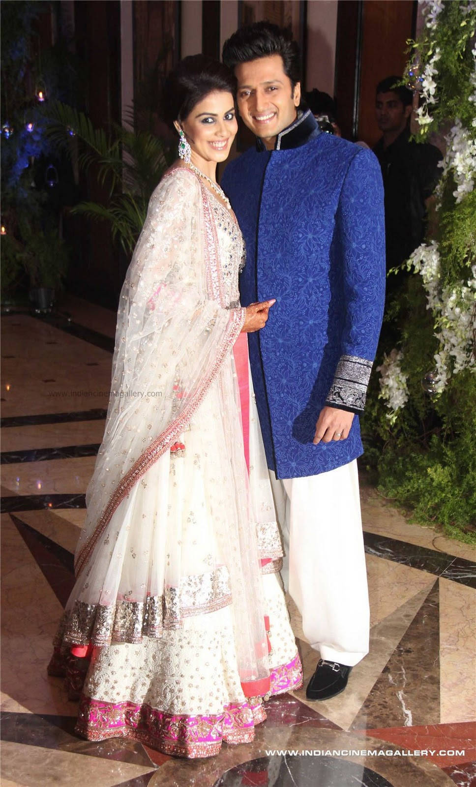 Riteish Deshmukh 與 Genelia 的溫馨時刻 壁紙 — 一對微笑的情侶穿著傳統印度服飾，在一個優雅的室內活動中拍照，場地有花卉裝飾和大理石地板。